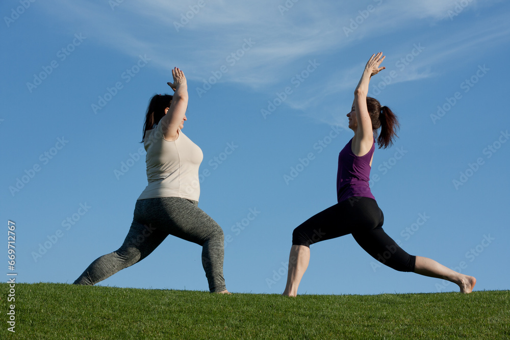 Two middle-aged women practice yoga in city park in warrior pose, blue sky background. Healthy lifestyle, fitness, Pilates, weight loss. overweight woman doing yoga with trainer. Two middle-aged women practice yoga in city park in warrior pose, blue sky background. Healthy lifestyle, fitness, Pilates, weight loss. overweight woman doing yoga with trainer.