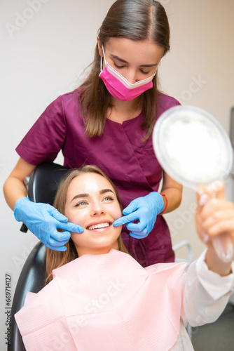 Young patient is looking in the mirror and admires her perfect smile after dental treatment.