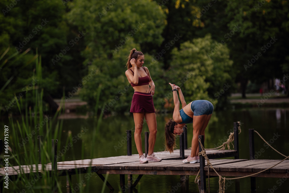 Fit, sporty females warm up and stretch in a green park. Picturesque bridge, water, and evening light create a healthy, attractive outdoor environment for exercising.
