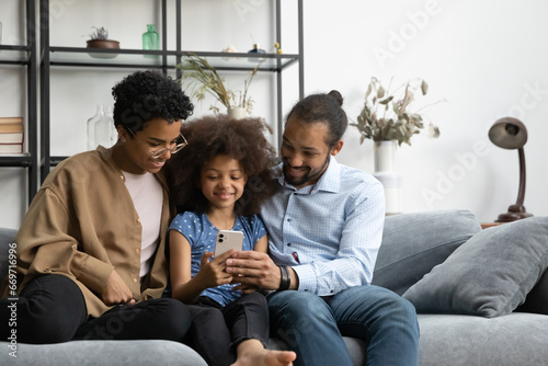 Happy young hipster Black parents and pre teen gen Z kid using online learning app on cellphone, relaxing on sofa at home. Mom, dad, daughter girl making video call on mobile phone, browsing Internet