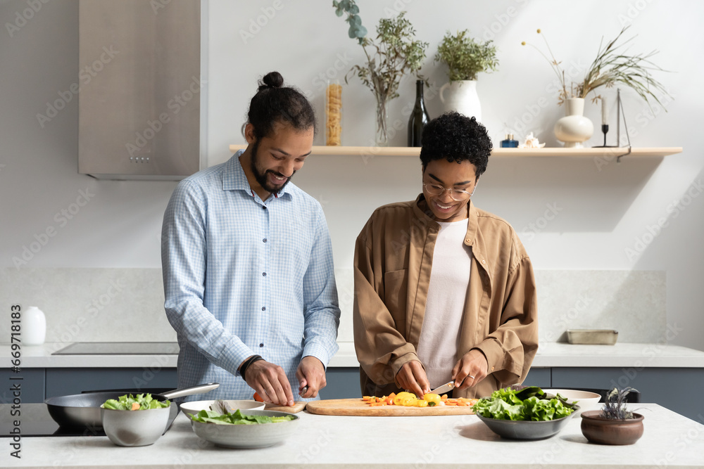 Happy young Black couple preparing homemade dinner in home kitchen, having fun, talking, smiling, laughing, enjoying date, cutting fresh vegetables for vegetarian salad together - obrazy, fototapety, plakaty 
