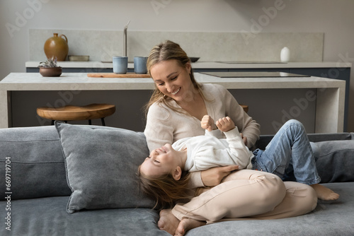 Excited happy mom tickling joyful adorable little daughter girl, playing with sweet child, relaxing on comfortable sofa at cozy home, enjoying family active games, motherhood, parenthood