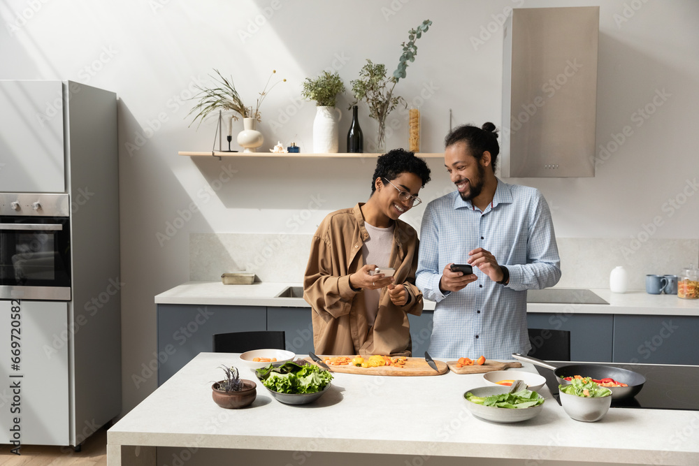 Joyful dating young African couple using online app on smartphones, cooking together in home kitchen, ordering food delivery on supermarket store internet website, consulting recipe on Internet Joyful dating young African couple using online app on smartphones, cooking together in home kitchen, ordering food delivery on supermarket store internet website, consulting recipe on Internet