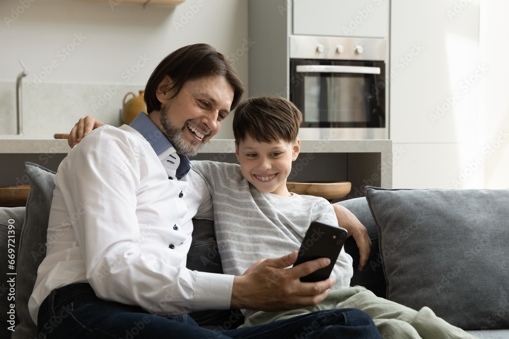 Excited joyful dad and happy teenage son kid sharing smartphone, talking on video call together, taking selfie on mobile phone. Father showing learning app, online shopping service to child Excited joyful dad and happy teenage son kid sharing smartphone, talking on video call together, taking selfie on mobile phone. Father showing learning app, online shopping service to child