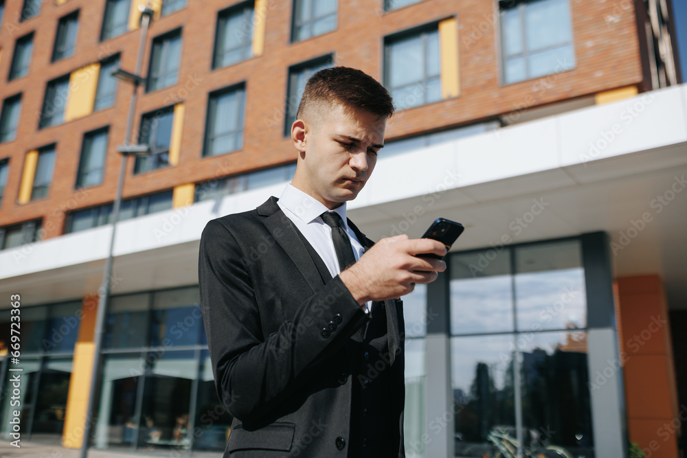 the business district, a young Caucasian American man in a well-fitted suit is absorbed in his smartphone, connecting to the internet. His confident posture highlights the role of modern technology