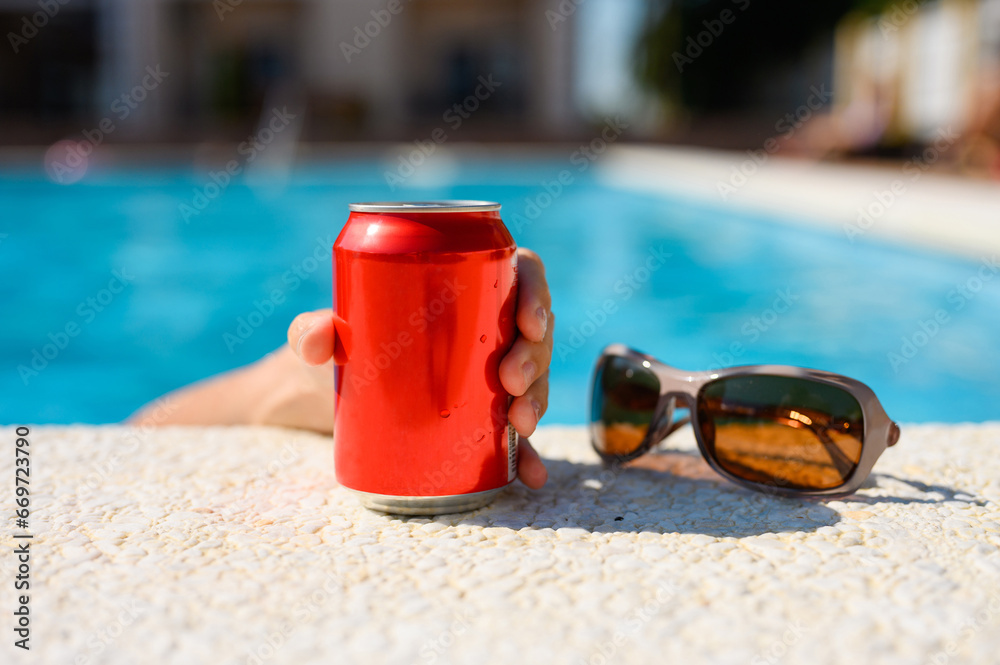 The hand of a vacationing tourist reaches for a red can of drink by the pool. Hot summer day by the pool. Close-up The hand of a vacationing tourist reaches for a red can of drink by the pool. Hot summer day by the pool. Close-up