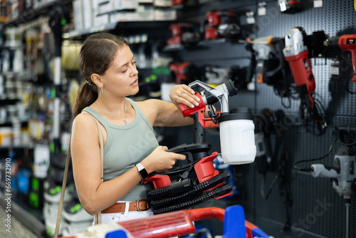 Girl buyer inspects modern power tool in construction hypermarket and selects picks up spray gun. Young woman near showcase with construction equipment