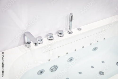 White acrylic bathtub with modern faucets and shower. The taps and mixer are made in chrome design. Water injectors at the bottom of the bathtub indicate the possibility of hydromassage