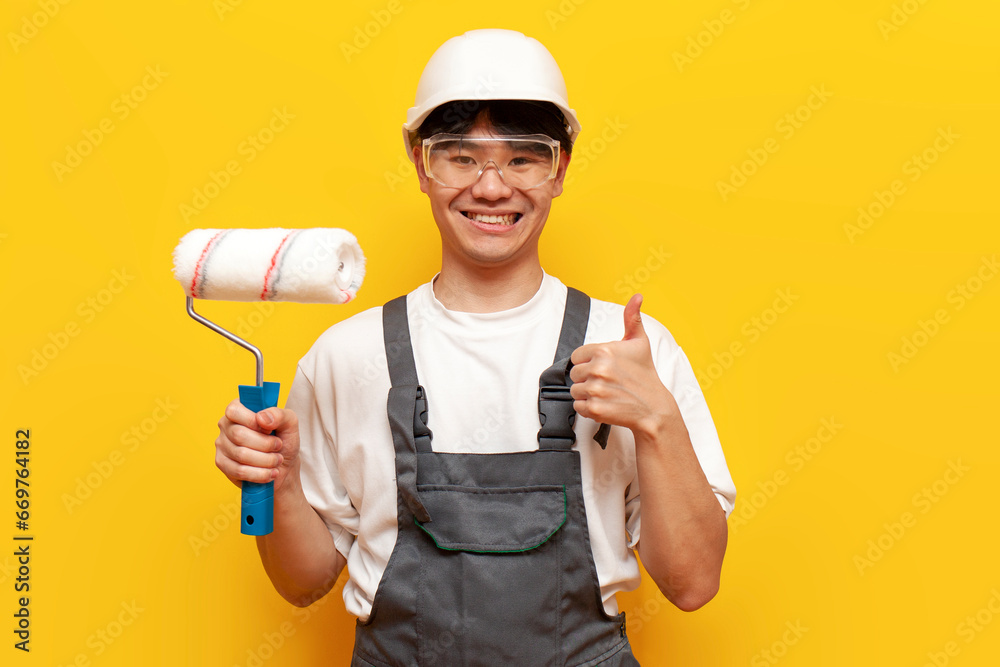 asian guy painter in uniform holds roller and shows like on yellow isolated background