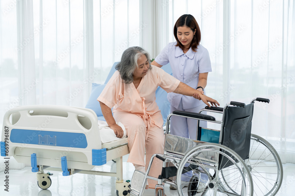 Obraz premium A nurse carries an elderly Asian patient into a wheelchair to take him to get an x-ray of his leg injured in an accident.