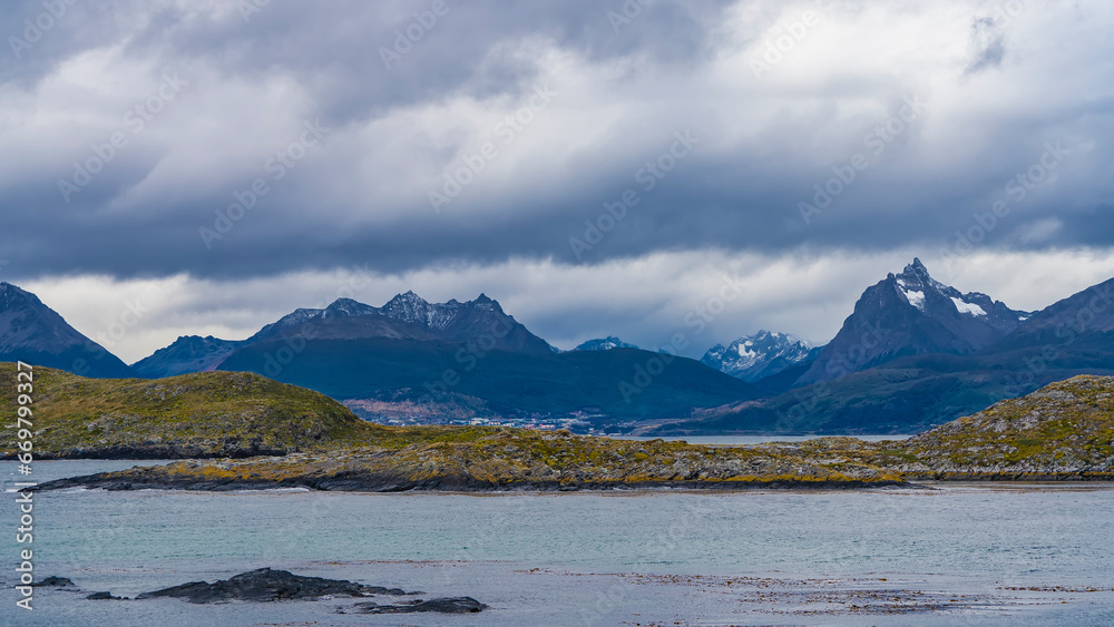 Picturesque mountain range of the Andes against a cloudy sky. The city houses of Ushuaia at the foot. In the foreground is the water of the Beagle Canal, an island with low-growing vegetation