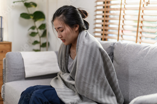 Young Asian woman sitting on couch have a cold and high fever have to rest at her home and absent from her job. Daily lifestyle health care concept.