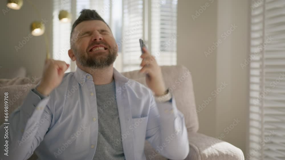 Portrait of overjoyed comic man screaming while watching results of sport game on smartphone. Excited hilarious guy sitting on sofa in living room. Bearded male clapping and screaming with excitement Portrait of overjoyed comic man screaming while watching results of sport game on smartphone. Excited hilarious guy sitting on sofa in living room. Bearded male clapping and screaming with excitement