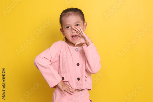 Cute attractive Asian little girl making announcement gesture over yellow background