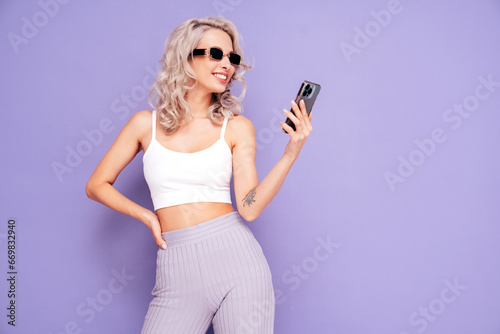 Young beautiful smiling female in trendy summer white top and pants clothes. Carefree blond woman posing near violet wall in studio. Positive model holds smartphone, uses phone apps, looks at screen