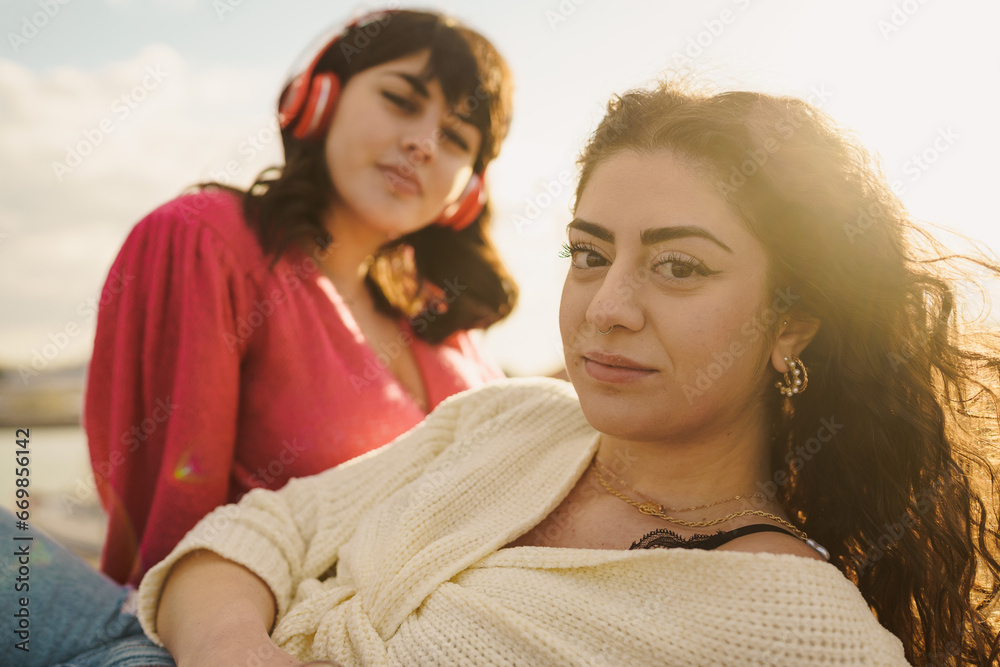 Two women, one wearing vibrant red with headphones and the other in an ivory sweater, share a tranquil moment by the sea, reflecting the warmth of the sun and the rhythm of the waves. Two women, one wearing vibrant red with headphones and the other in an ivory sweater, share a tranquil moment by the sea, reflecting the warmth of the sun and the rhythm of the waves.