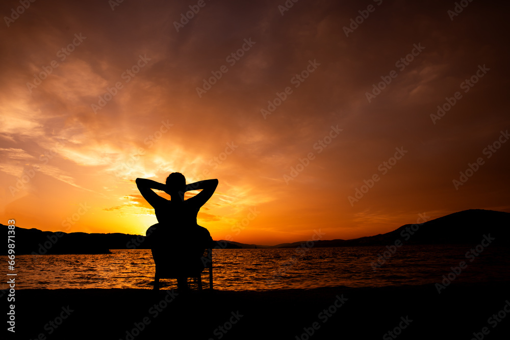sylwetka osoby o zachodzie słońca siedząca i podziwiająca zachód słońca, relaks nad morzem - obrazy, fototapety, plakaty 