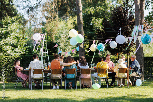 Family and friends sitting at the party table during a summer garden party outdoors. Rear view of people sitting at the table.