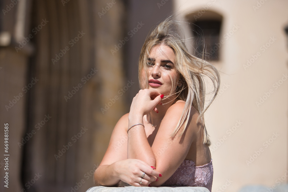 Portrait of young woman, blonde, green eyes, with pink top, leaning on a stone column, letting the wind move her hair. Concept beauty, hairstyles, wind, mane. Portrait of young woman, blonde, green eyes, with pink top, leaning on a stone column, letting the wind move her hair. Concept beauty, hairstyles, wind, mane.