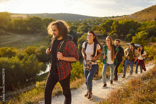 Group of people hiking or trekking together. Team of young male and female hikers enjoying good sunny day, carrying backpacks and going along hill track, with beautiful rural landscape in background