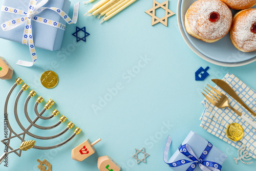 Jewish Hanukkah table setup idea. Top view of plate with traditional donuts, napkin, cutlery, Star of David signs, gifts, menorah, gelt, dreidel on soft blue backdrop with frame for text or marketing