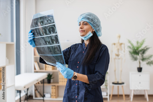 Medical female specialist in protective scrubs and cap holding x-ray of patient's brain and looking at it. Confident indian doctor inspecting roentgen in her comfortable light office. photo