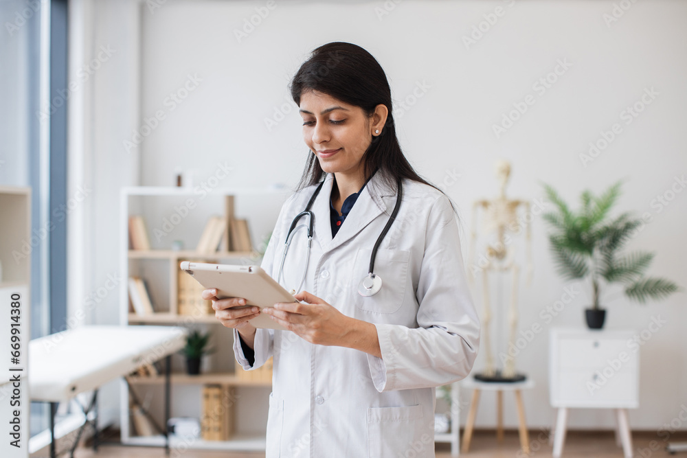 Focused hindu female in white lab coat holding tablet while posing on background of anatomical model in doctor's office. Concept of people, medicine and technology. Focused hindu female in white lab coat holding tablet while posing on background of anatomical model in doctor's office. Concept of people, medicine and technology.