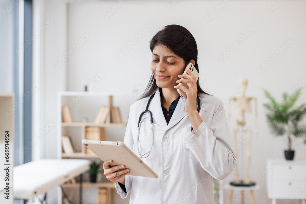 Front view of female physician standing and talking on mobile phone while looking on tablet screen. Competent hindu doctor using modern devices for work and consultation from hospital.
