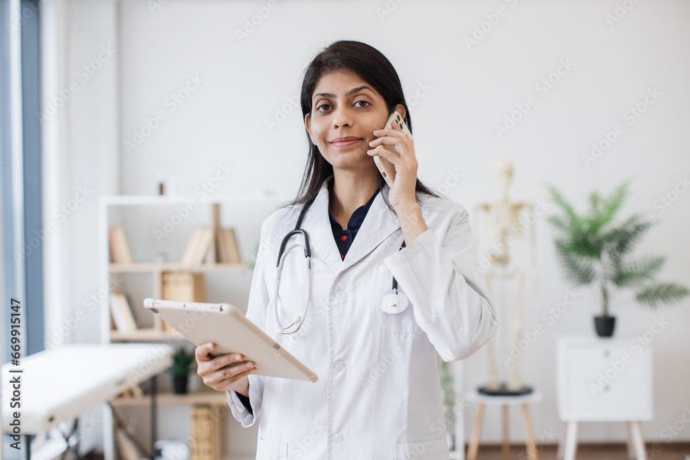 Front view of female physician standing and talking on mobile phone while looking on tablet screen. Competent hindu doctor using modern devices for work and consultation from hospital. Front view of female physician standing and talking on mobile phone while looking on tablet screen. Competent hindu doctor using modern devices for work and consultation from hospital.