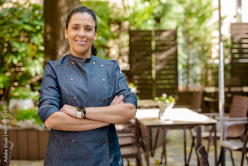 Female chef wears blue coat, happy smiling poses with arms crossed outdoors on the terrace of a restaurant. Restaurant worker, culinary gourmet, pastry chef. photo