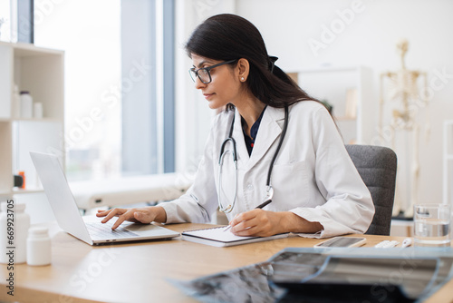 Side view of indian female doctor using portable computer while sitting at office desk in health center interior. Efficient mature general practitioner checking patient records via gadget.