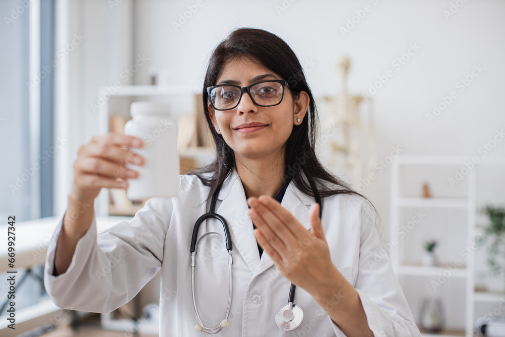 Professional therapist in lab coat and glasses standing with white pill bottle in hands. Successful indian doctor in eyeglasses displaying most effective drugs for treatment on workplace background.