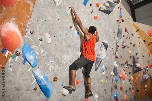 athletic african american man in orange shirt gripping on boulders while climbing up rock wall