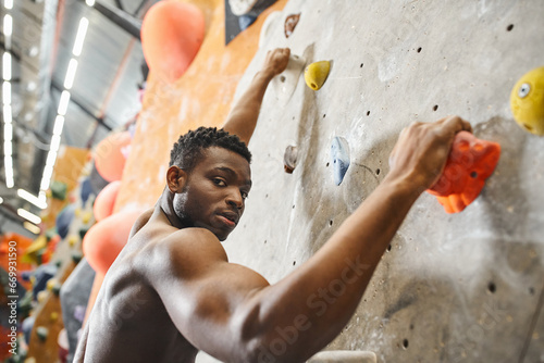 good looking shirtless african american man gripping on boulders and looking straight at camera