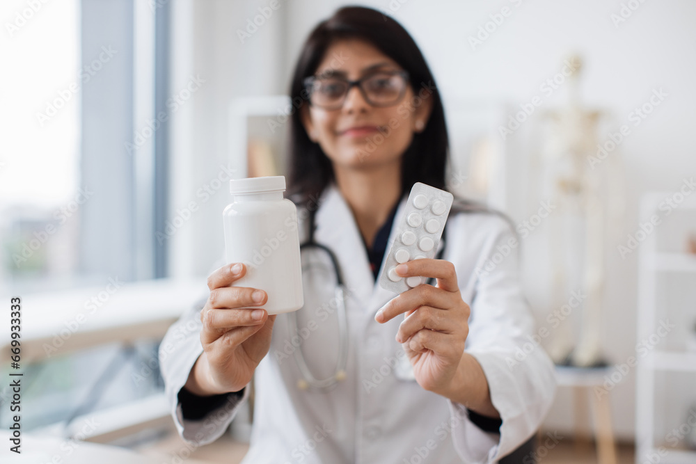 Friendly female therapist in glasses showing white jar of medicine while sitting at desk. Experienced indian woman in white coat recommending best alternative of treating at doctor's office. Friendly female therapist in glasses showing white jar of medicine while sitting at desk. Experienced indian woman in white coat recommending best alternative of treating at doctor's office.