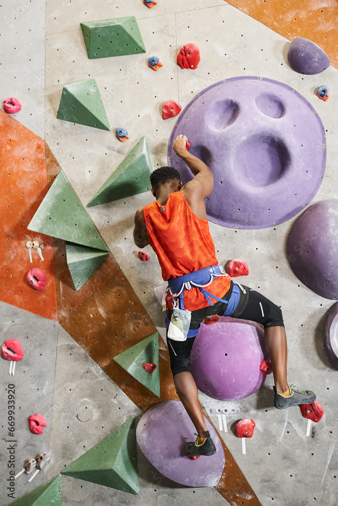 vertical shot of muscular african american man with alpine harness climbing up rock wall, bouldering