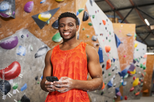 jolly african american man in orange shirt holding mobile phone and looking at camera, rock climbing © LIGHTFIELD STUDIOS