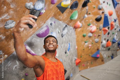 happy african american man taking selfie and smiling cheerfully with climbing wall backdrop