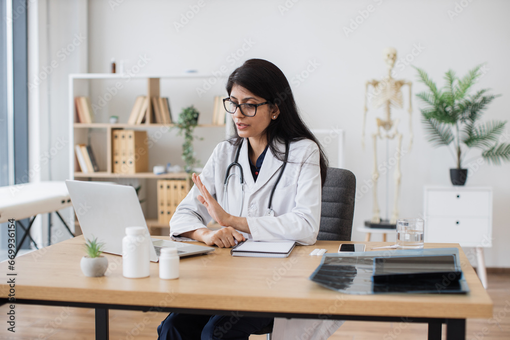 Mature indian woman in lab coat and glasses waving hello to ill patient while having conversation via computer in office interior. Family doctor using modern technologies to make treatment plan. Mature indian woman in lab coat and glasses waving hello to ill patient while having conversation via computer in office interior. Family doctor using modern technologies to make treatment plan.