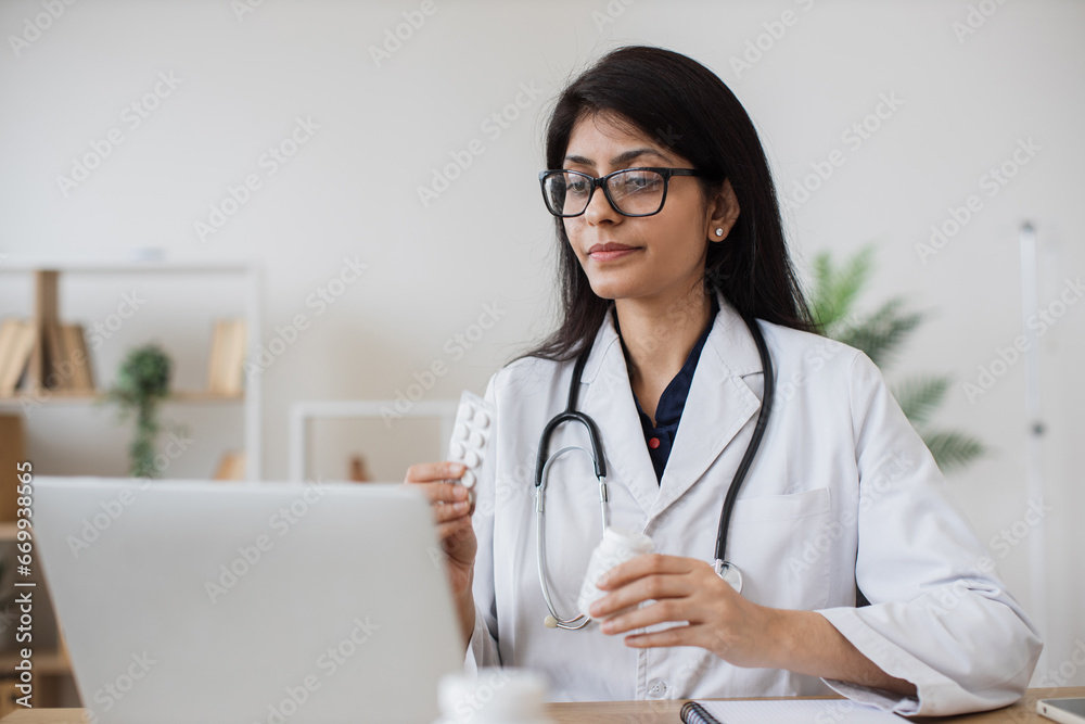 Friendly woman sitting at table and having conversation with patient on video call while holding medicine in hands. Hindu therapist recommending means of treatment using modern laptop in office. Friendly woman sitting at table and having conversation with patient on video call while holding medicine in hands. Hindu therapist recommending means of treatment using modern laptop in office.