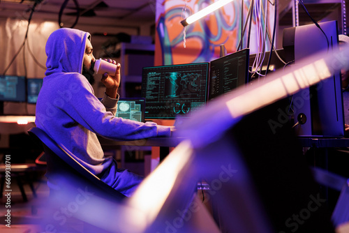 Hacker drinking coffee and doing illegal activities on computer late at night. Criminal holding beverage to go while cracking password and using malicious software in abandoned warehouse