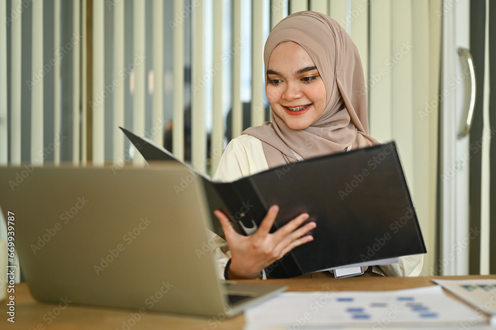 Fototapeta premium Smiling Muslim businesswoman reading document, checking financial report at office desk