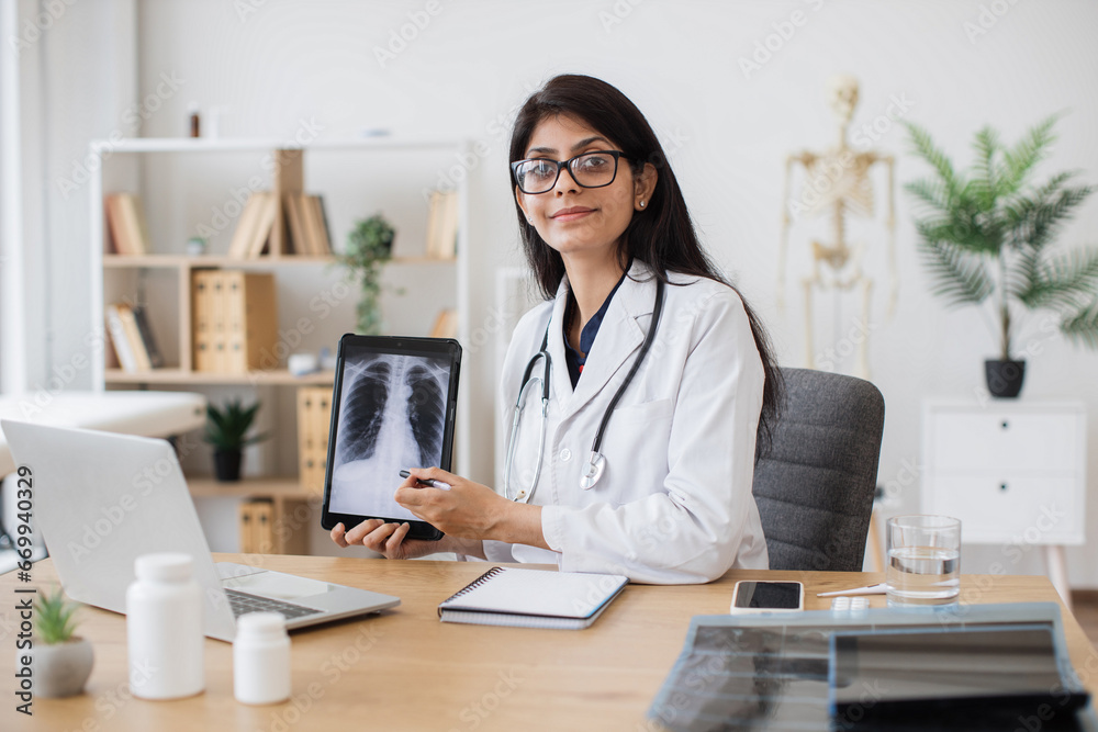 Concentrated female doctor talking on video call while showing result of examination of patient with tuberculosis. Indian lady consulting person using laptop and tablet in spacious clinic room. Concentrated female doctor talking on video call while showing result of examination of patient with tuberculosis. Indian lady consulting person using laptop and tablet in spacious clinic room.