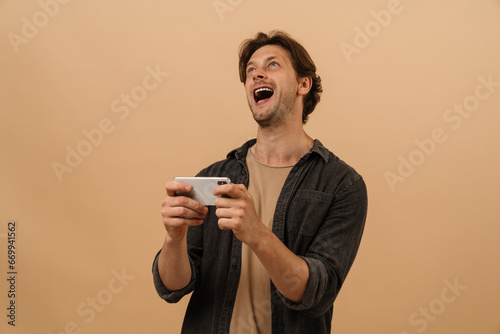 Excited man playing game with smartphone while standing isolated over beige wall © Drobot Dean