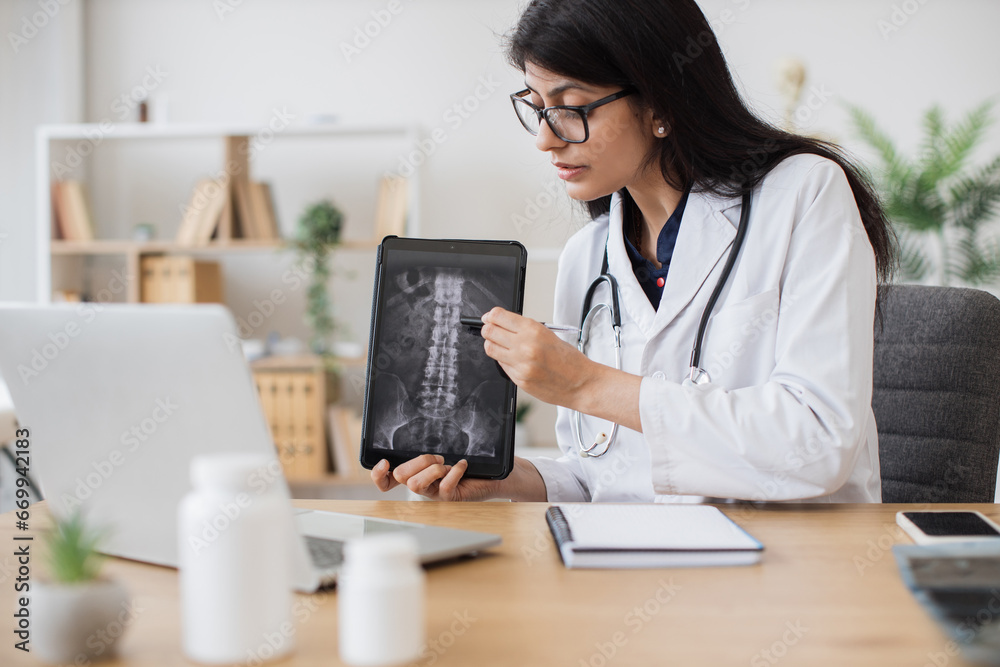 Indian female specialist sitting in front of digital laptop and consulting patient with spinal disease. Focused nurse using tablet with x-ray scan to showing problem area in medical facility. Indian female specialist sitting in front of digital laptop and consulting patient with spinal disease. Focused nurse using tablet with x-ray scan to showing problem area in medical facility.