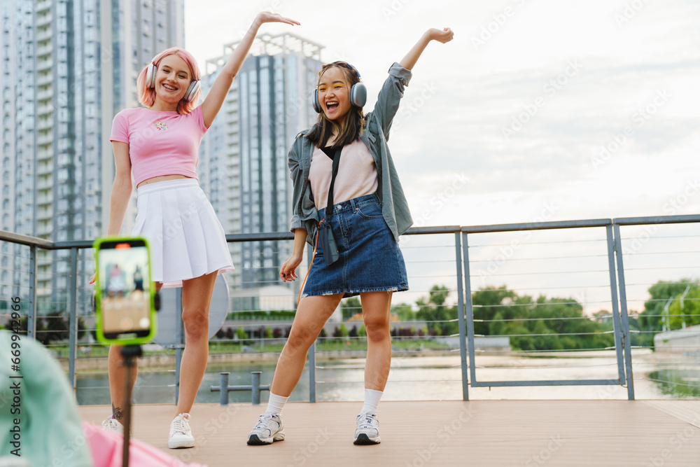 Obraz premium Two joyful girls making dancing videos with smartphone camera on city waterfront