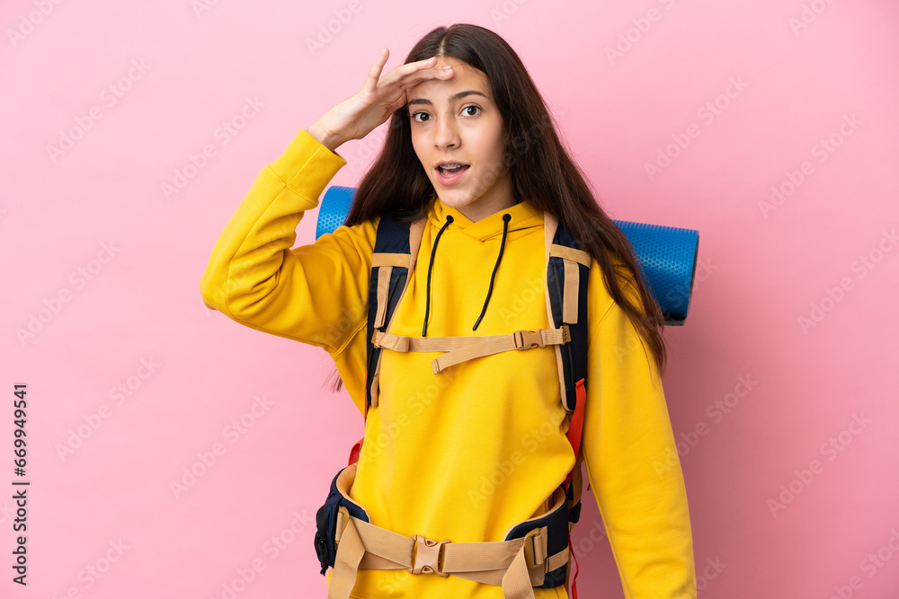 Young mountaineer girl with a big backpack isolated on pink background doing surprise gesture while looking to the side Young mountaineer girl with a big backpack isolated on pink background doing surprise gesture while looking to the side