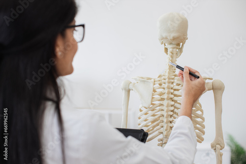 View from shoulder of indian female holding personal tablet while touching with pen backbone of modern human body model. Young intern carefully examining skeleton and studying spine problems. photo