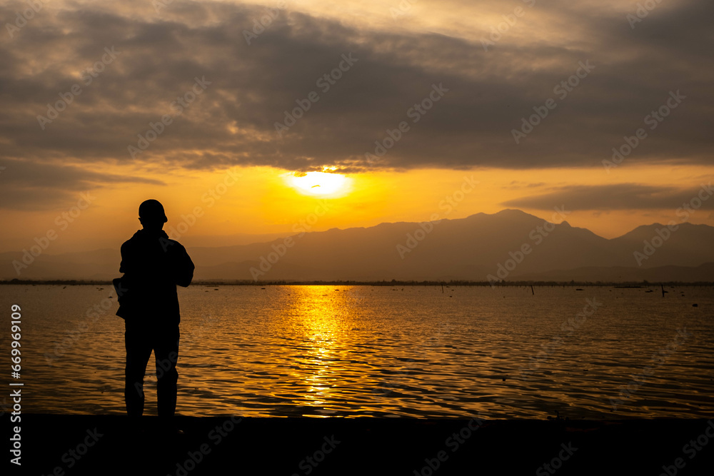 A young man in black is taking pictures of the sunset behind the mountain. And there are bright colored lights reflected on the water surface as a beautiful background. A young man in black is taking pictures of the sunset behind the mountain. And there are bright colored lights reflected on the water surface as a beautiful background.