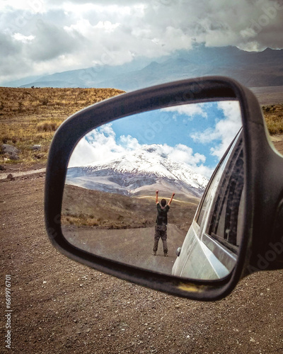 homem de braçso erguidos com imagem refletida no retrovisor do carro com vuclão cotopaxi ao fundo 